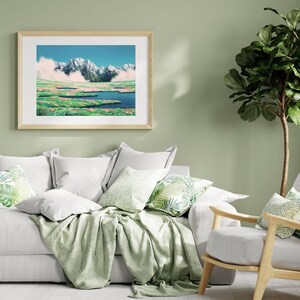 Pu&ograve; includere: Una stampa artistica incorniciata raffigura un paesaggio montano con cime innevate, un lago blu e campi verdi. L'opera d'arte &egrave; esposta sopra un divano bianco con cuscini decorativi e una coperta verde. Una pianta verde &egrave; sullo sfondo.