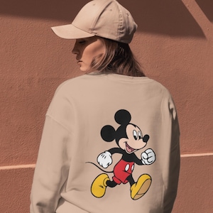 May include: A beige crewneck sweatshirt featuring a graphic print of Mickey Mouse on the back, depicted in a running pose with black ears, red shorts, and yellow shoes. The garment is paired with a matching beige baseball cap worn on the head.
