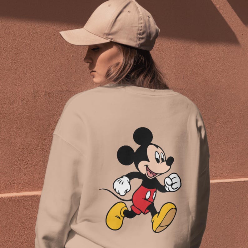 May include: A beige crewneck sweatshirt featuring a graphic print of Mickey Mouse on the back, depicted in a running pose with black ears, red shorts, and yellow shoes. The garment is paired with a matching beige baseball cap worn on the head.