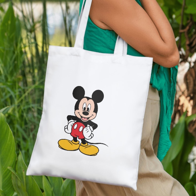 May include: A white tote bag featuring a centered graphic of Mickey Mouse. The character is depicted with his signature black ears, red shorts, white gloves, and large yellow shoes, standing with hands on his hips against the plain white fabric.