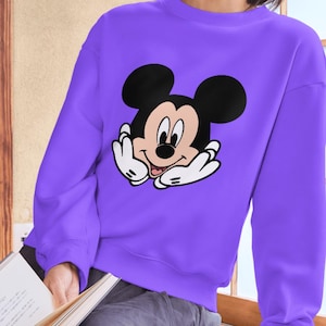May include: A vibrant purple long-sleeve sweatshirt featuring a large, centered graphic of Mickey Mouse. The character is depicted with a black head, large ears, and white gloved hands resting against the face, showcasing a cheerful expression.