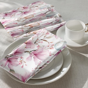 Può includere: Tazza e piattino in ceramica bianca con manico dorato, su un piatto bianco. Due tovaglioli bianchi piegati con un motivo floreale rosa e accenti di uova azzurre sono su un piatto bianco. I tovaglioli presentano fiori di magnolia rosa e rami marroni.
