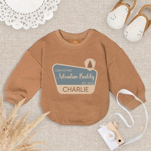 May include: A tan baby onesie with long sleeves. The front features a graphic that reads "OUR FUTURE Adventure Buddy EST. 2025 CHARLIE" in blue and tan. A pair of white and brown baby shoes and a toy camera are also in the image.