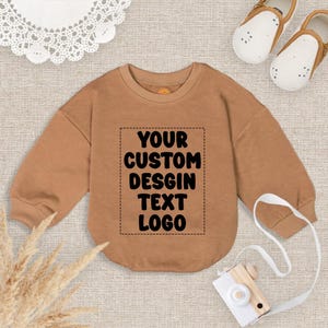 May include: A tan-colored baby bodysuit with long sleeves. The front of the bodysuit features a rectangular area with the text "YOUR CUSTOM DESIGN TEXT LOGO" in black. The image also includes a pair of baby shoes and a toy camera.
