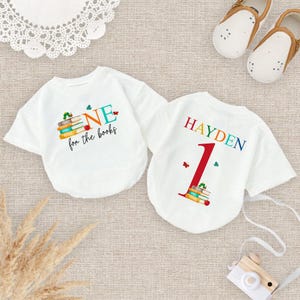 May include: White baby bodysuit with the text "ONE for the books" and a stack of books graphic. The back of the bodysuit has the name "HAYDEN" and the number "1" with a stack of books graphic.