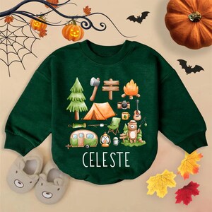 May include: A dark green sweatshirt featuring a camping-themed graphic with a tent, campfire, and bear. The name "CELESTE" is printed in white. The image is decorated with Halloween elements like pumpkins, bats, and leaves.