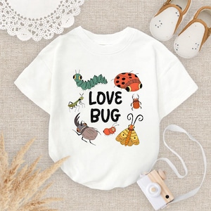 May include: White short-sleeved t-shirt with a colorful bug graphic. The design includes a green caterpillar, a red ladybug, and the text "LOVE BUG". A toy camera and baby shoes are in the background.