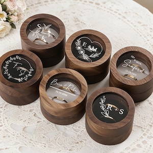 以下が含まれることがあります： 透明な蓋が付いた丸い木製のリングボックスが6つ。それぞれに金の指輪が入っています。ボックスにはイニシャルと日付が刻印されています。イニシャルの周りには花と葉のデザインがあります。背景には花のブーケがあります。