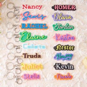 Puede incluir: Una colección de llaveros con nombres coloridos con llaveros plateados. Los nombres incluyen "Nancy", "Sevi", "Rachel", "Elaine", "Calista", "Truda", "Juliet", "Stella", "Homer", "Olivia", "Jordan", "Kirsten", "Nestor", "Harvey", "Kevin" y "Nicole".
