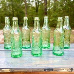 Group of seven aqua glass 3 Centa soda bottles arranged outdoors on a white surface.