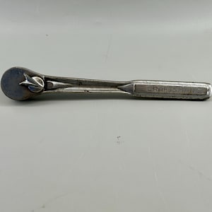 May include: A silver-colored metal Craftsman ratchet tool featuring a circular head with a central adjustment switch and a long, straight handle with the brand name Craftsman embossed clearly on the side of the metal shaft.
