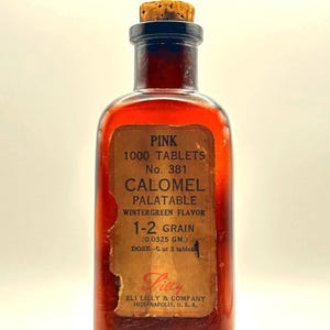 May include: A vintage amber glass bottle with a cork stopper. The bottle has a paper label with the text "PINK CALOMEL" and other details. The label is aged and slightly torn, adding to its antique appearance. The bottle is likely from the early 20th century.