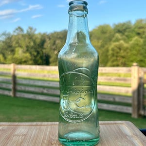 May include: A clear, light green glass bottle with an embossed circular design on the front. The text inside the circle reads The Original 5 Cent Drink. The bottle has a narrow neck and a rounded rim, featuring a smooth, transparent glass surface.