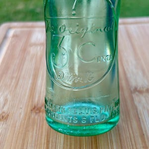 May include: A vintage light green glass bottle featuring an embossed circular logo with the text The Original 3 Cents Drink. The base of the bottle includes embossed text reading REG U.S. PAT. OFF. and CONTENTS 6 FLUID OZS.