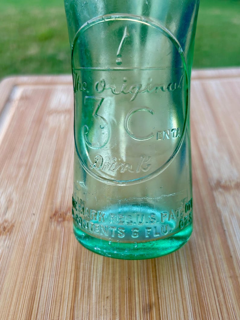 May include: A vintage light green glass bottle featuring an embossed circular logo with the text The Original 3 Cents Drink. The base of the bottle includes embossed text reading REG U.S. PAT. OFF. and CONTENTS 6 FLUID OZS.