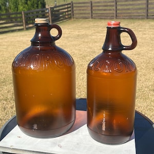 May include: Two brown glass jugs with handles and metal caps. The word "CLOROX" is embossed on the front of each jug. The jugs are sitting on a round, dark-colored table outside on a sunny day.