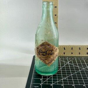 Pode incluir: Uma garrafa de vidro verde-claro vintage da Coca-Cola com um r&oacute;tulo de papel em forma de diamante escrito Coca-Cola, Atlanta, GA. Uma r&eacute;gua vertical mostra uma altura de 19 cm e uma r&eacute;gua horizontal mostra uma base de 6,4 cm.