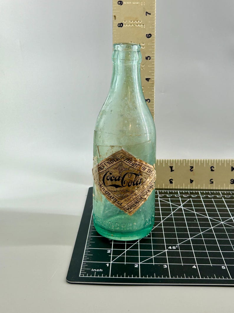 Pode incluir: Uma garrafa de vidro verde-claro vintage da Coca-Cola com um r&oacute;tulo de papel em forma de diamante escrito Coca-Cola, Atlanta, GA. Uma r&eacute;gua vertical mostra uma altura de 19 cm e uma r&eacute;gua horizontal mostra uma base de 6,4 cm.