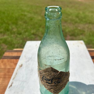 Pode incluir: Uma garrafa de Coca-Cola vintage de vidro verde claro com gargalo estreito e borda arredondada. Um r&oacute;tulo de papel desgastado em forma de diamante est&aacute; colado na parte inferior, exibindo o logotipo da Coca-Cola e texto parcialmente vis&iacute;vel.