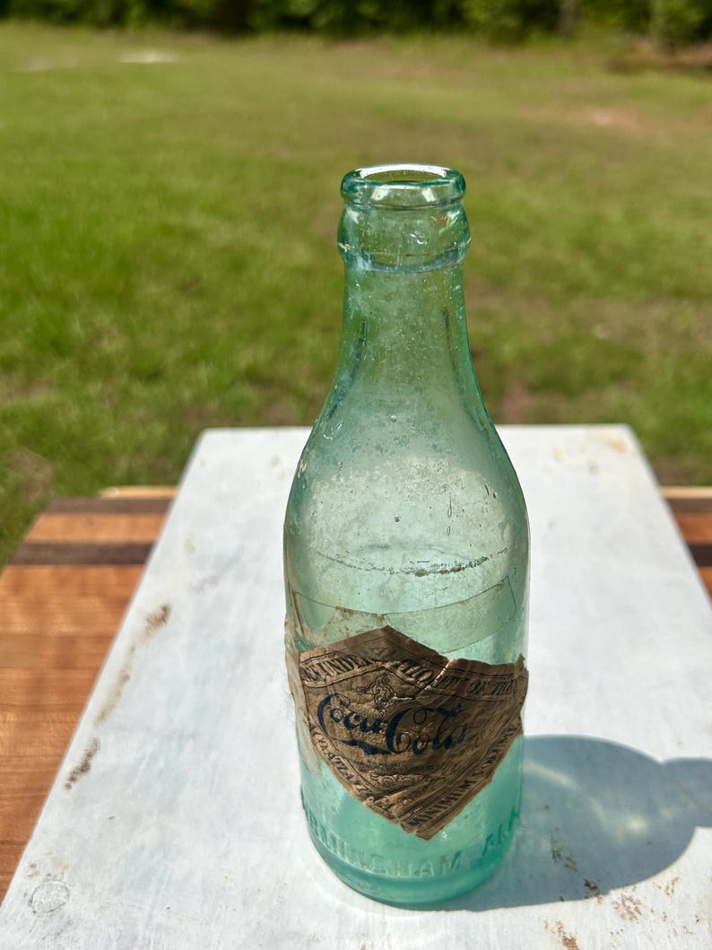 Pode incluir: Uma garrafa de Coca-Cola vintage de vidro verde claro com gargalo estreito e borda arredondada. Um r&oacute;tulo de papel desgastado em forma de diamante est&aacute; colado na parte inferior, exibindo o logotipo da Coca-Cola e texto parcialmente vis&iacute;vel.