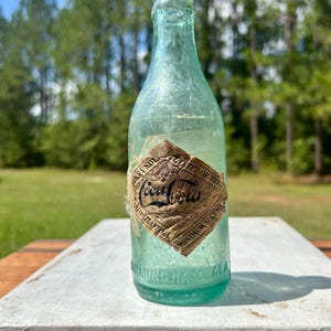 Pode incluir: Uma garrafa de vidro verde-claro vintage da Coca-Cola com um r&oacute;tulo de papel desgastado em forma de diamante. O r&oacute;tulo apresenta o log&oacute;tipo da Coca-Cola, o texto "Minimum Volume" e "Atlanta, GA". A base tem "Birmingham, Ala." gravado.