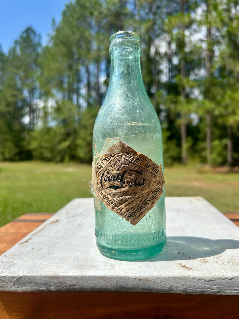 Pode incluir: Uma garrafa de vidro verde-claro vintage da Coca-Cola com um r&oacute;tulo de papel desgastado em forma de diamante. O r&oacute;tulo apresenta o log&oacute;tipo da Coca-Cola, o texto "Minimum Volume" e "Atlanta, GA". A base tem "Birmingham, Ala." gravado.