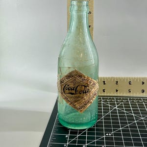 Pode incluir: Uma garrafa de vidro verde-claro vintage da Coca-Cola com um r&oacute;tulo de papel em forma de diamante com os dizeres Coca-Cola, Atlanta GA e Minimum Volume. Uma r&eacute;gua de madeira indica uma altura de aproximadamente 19,7 cm.