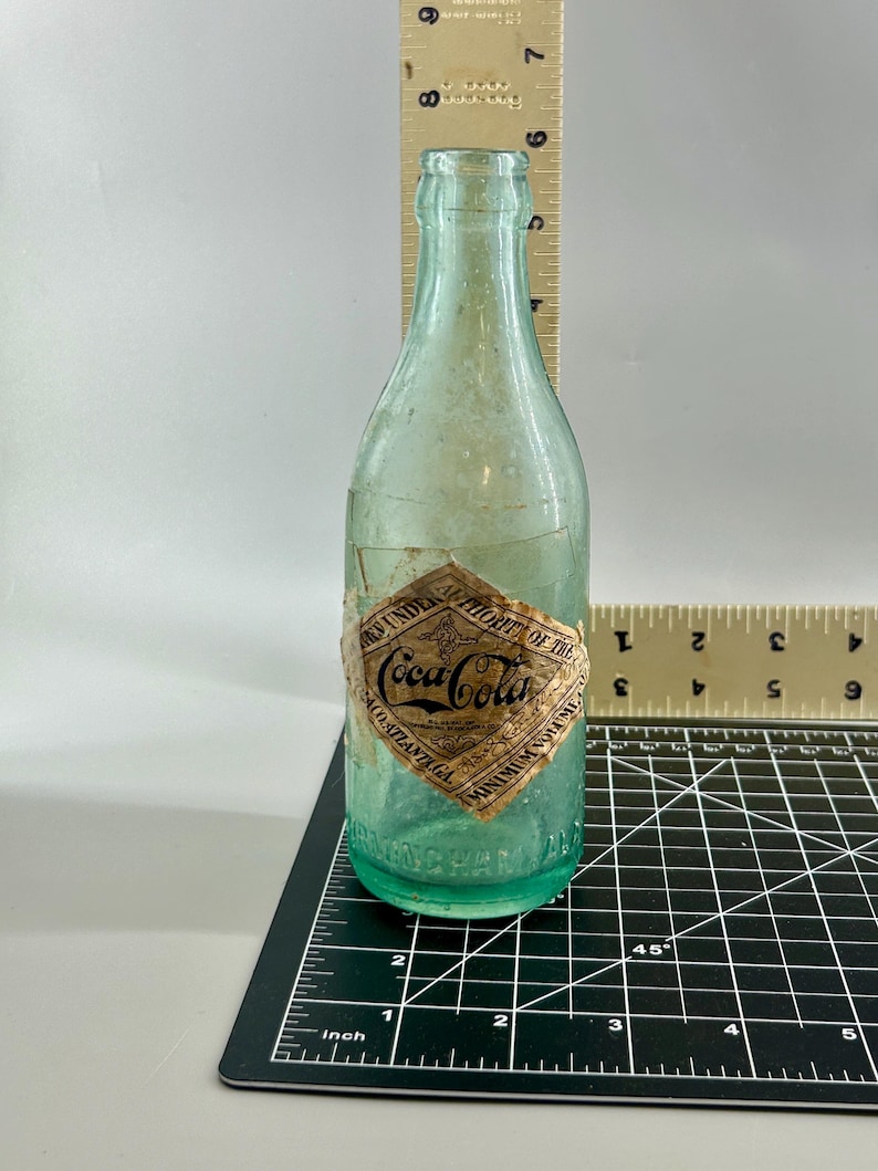 Pode incluir: Uma garrafa de vidro verde-claro vintage da Coca-Cola com um r&oacute;tulo de papel em forma de diamante com os dizeres Coca-Cola, Atlanta GA e Minimum Volume. Uma r&eacute;gua de madeira indica uma altura de aproximadamente 19,7 cm.