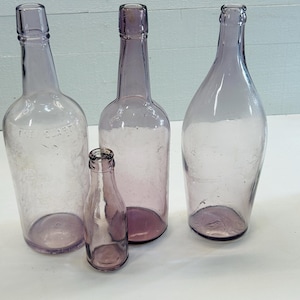 May include: Four antique, clear glass bottles in varying sizes and shapes. The bottles are a soft purple hue, with visible imperfections and signs of age. One bottle has the text "THE CLART" embossed on its side. The bottles are arranged on a white surface.