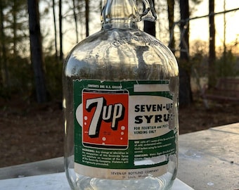May include: A clear glass 7-Up syrup bottle with a handle and a gray cap. The label features the 7-Up logo in red and white, with text indicating it's for fountain and post-vending use. The bottle holds one U.S. gallon.