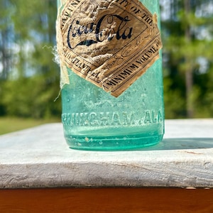 Pode incluir: Uma garrafa de Coca-Cola de vidro verde claro vintage com um r&oacute;tulo de papel desgastado em forma de diamante com texto preto. O r&oacute;tulo exibe o logotipo da Coca-Cola, enquanto a base de vidro tem gravado o texto BIRMINGHAM ALA em relevo.