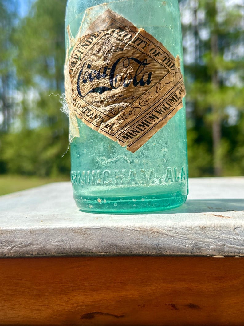 Pode incluir: Uma garrafa de Coca-Cola de vidro verde claro vintage com um r&oacute;tulo de papel desgastado em forma de diamante com texto preto. O r&oacute;tulo exibe o logotipo da Coca-Cola, enquanto a base de vidro tem gravado o texto BIRMINGHAM ALA em relevo.