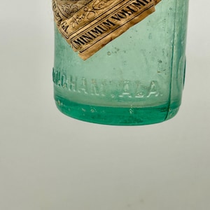 Pode incluir: Uma garrafa de vidro verde claro vintage com um r&oacute;tulo de papel parcialmente rasgado com o texto Coca-Cola e Minimum Volume 6 OZ. Na superf&iacute;cie inferior do vidro est&aacute; gravado o texto BIRMINGHAM, ALA. A garrafa tem formato cil&iacute;ndrico.