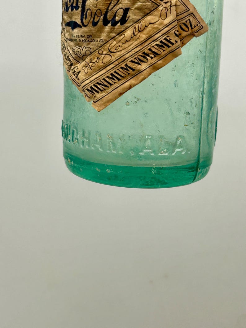 Pode incluir: Uma garrafa de vidro verde claro vintage com um r&oacute;tulo de papel parcialmente rasgado com o texto Coca-Cola e Minimum Volume 6 OZ. Na superf&iacute;cie inferior do vidro est&aacute; gravado o texto BIRMINGHAM, ALA. A garrafa tem formato cil&iacute;ndrico.