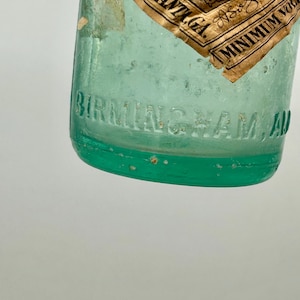 Pode incluir: Uma garrafa de vidro verde claro com uma &aacute;rea de r&oacute;tulo em relevo com o texto BIRMINGHAM, ALA. Um r&oacute;tulo de papel castanho parcial est&aacute; anexado, exibindo o texto COCA-COLA, ATLANTA, GA., REG. U.S. PAT. OFF., COPYRIGHT 1907, BY COCA-COLA CO. e MINIMUM VOL.