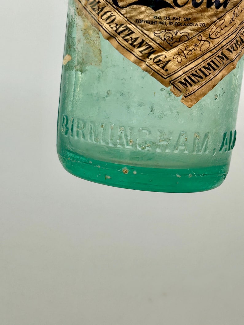 Pode incluir: Uma garrafa de vidro verde claro com uma &aacute;rea de r&oacute;tulo em relevo com o texto BIRMINGHAM, ALA. Um r&oacute;tulo de papel castanho parcial est&aacute; anexado, exibindo o texto COCA-COLA, ATLANTA, GA., REG. U.S. PAT. OFF., COPYRIGHT 1907, BY COCA-COLA CO. e MINIMUM VOL.