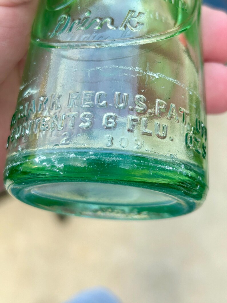 May include: A light green glass bottle with embossed text that reads Drink, Trade Mark Reg. U.S. Pat. Off., and Contents 6 Flu. Ozs. The bottle features a cylindrical base and a curved neck, with the number 309 visible near the bottom edge.