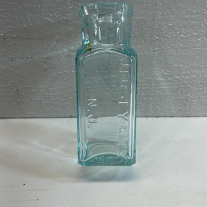 May include: A rectangular, light blue glass bottle with the text "JERSEY CITY N.J." embossed on the front. The bottle has a square base and a short neck. The glass has a slightly aged appearance.