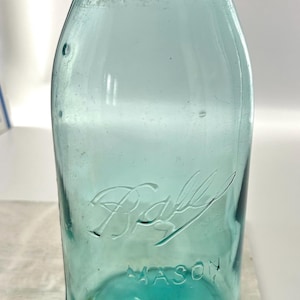 May include: A vintage aqua-colored Ball Mason jar with a dark metal lid. The jar has embossed lettering that reads "Ball" and "Mason". The glass jar is a classic design, often used for canning and preserving food.