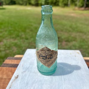 Pode incluir: Uma garrafa de vidro verde-claro vintage da Coca-Cola com um r&oacute;tulo de papel em forma de diamante. O r&oacute;tulo apresenta o log&oacute;tipo da Coca-Cola, o texto "Atlanta, GA" e "Minimum Volume 6 Fl. Oz.". A garrafa tem um gargalo estreito.