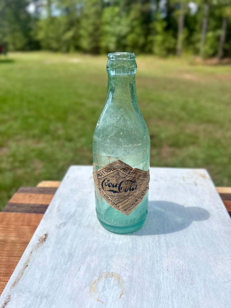 Pode incluir: Uma garrafa de vidro verde-claro vintage da Coca-Cola com um r&oacute;tulo de papel em forma de diamante. O r&oacute;tulo apresenta o log&oacute;tipo da Coca-Cola, o texto "Atlanta, GA" e "Minimum Volume 6 Fl. Oz.". A garrafa tem um gargalo estreito.
