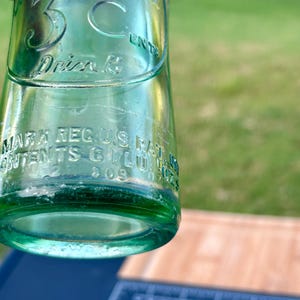 May include: A vintage green glass soda bottle featuring embossed text that reads The Original 3 Cent Drink, along with Mark Reg U.S. Pat. Off. and Contents 6 Fl. Oz. The bottle has a cylindrical body with a smooth surface and clear glass construction.