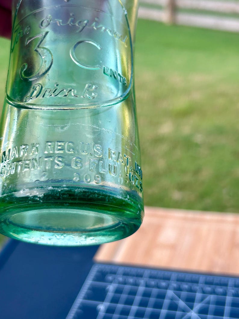 May include: A vintage green glass soda bottle featuring embossed text that reads The Original 3 Cent Drink, along with Mark Reg U.S. Pat. Off. and Contents 6 Fl. Oz. The bottle has a cylindrical body with a smooth surface and clear glass construction.