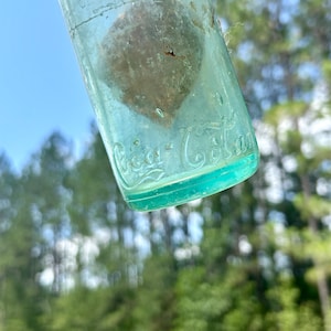Pode incluir: Uma garrafa de vidro verde claro vintage com uma superf&iacute;cie desgastada e um interior texturizado. O vidro apresenta o log&oacute;tipo da Coca-Cola em relevo em letra cursiva perto da base. A garrafa &eacute; transl&uacute;cida e mostra sinais de idade.
