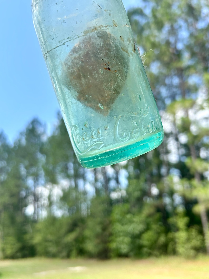 Pode incluir: Uma garrafa de vidro verde claro vintage com uma superf&iacute;cie desgastada e um interior texturizado. O vidro apresenta o log&oacute;tipo da Coca-Cola em relevo em letra cursiva perto da base. A garrafa &eacute; transl&uacute;cida e mostra sinais de idade.
