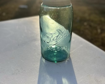 Puede incluir: Un frasco Mason de vidrio azul claro vintage con una tapa de rosca blanca. El vidrio presenta el texto en relieve Ball en cursiva y la palabra MASON en letras mayúsculas en la superficie frontal. El frasco tiene un cuerpo cilíndrico.