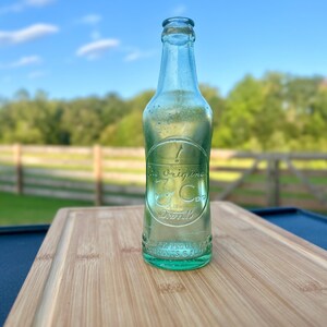 May include: A vintage light green glass soda bottle with an embossed circular logo featuring the text 'The original 3 Cents Drink' and additional embossed lettering near the base, including 'REG. U.S. PAT. OFF.' and 'CONTENTS 6 FLUID OUNCES'.