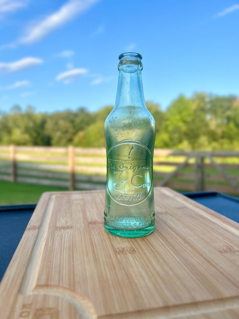 May include: A vintage light green glass soda bottle with an embossed circular logo featuring the text 'The original 3 Cents Drink' and additional embossed lettering near the base, including 'REG. U.S. PAT. OFF.' and 'CONTENTS 6 FLUID OUNCES'.