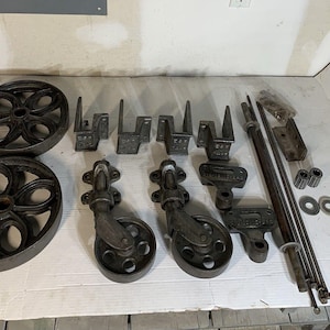May include: A collection of antique cast iron hardware components, including two large wheels, casters, brackets, and rods. The pieces are dark gray and appear to be salvaged or repurposed. Ideal for restoration projects or industrial decor.