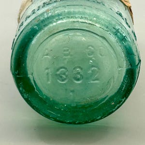 Pode incluir: Uma garrafa de vidro verde-claro vintage com um r&oacute;tulo de papel bege parcial enrolado no gargalo. A base circular apresenta um texto em relevo com A B CO e o n&uacute;mero 1332. O vidro &eacute; transl&uacute;cido com bolhas de ar vis&iacute;veis no material.